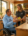 A man in a wheelchair wearing a blue hoodie handing a bowl of fruit to his wife in a cozy kitchen setting to represent the Wear the Message collection.